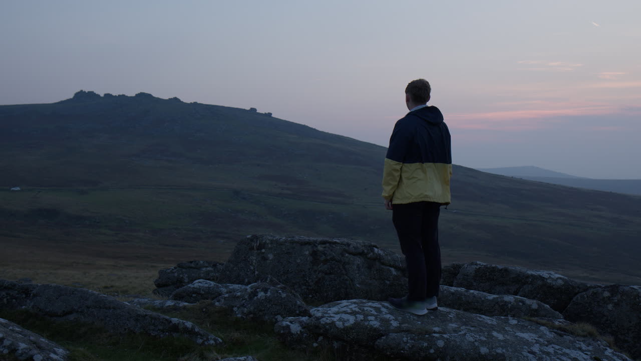 Dartmoor, Devon, Inghilterra, Regno Unito. Un uomo sta in piedi su una collina rocciosa, osservando il paesaggio sereno e vasto immerso nelle calde tonalità del tramonto. Piano medio.