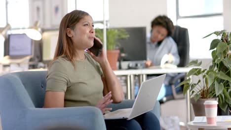 mujer de negocios casual biracial hablando en teléfono inteligente y usando computadora portátil