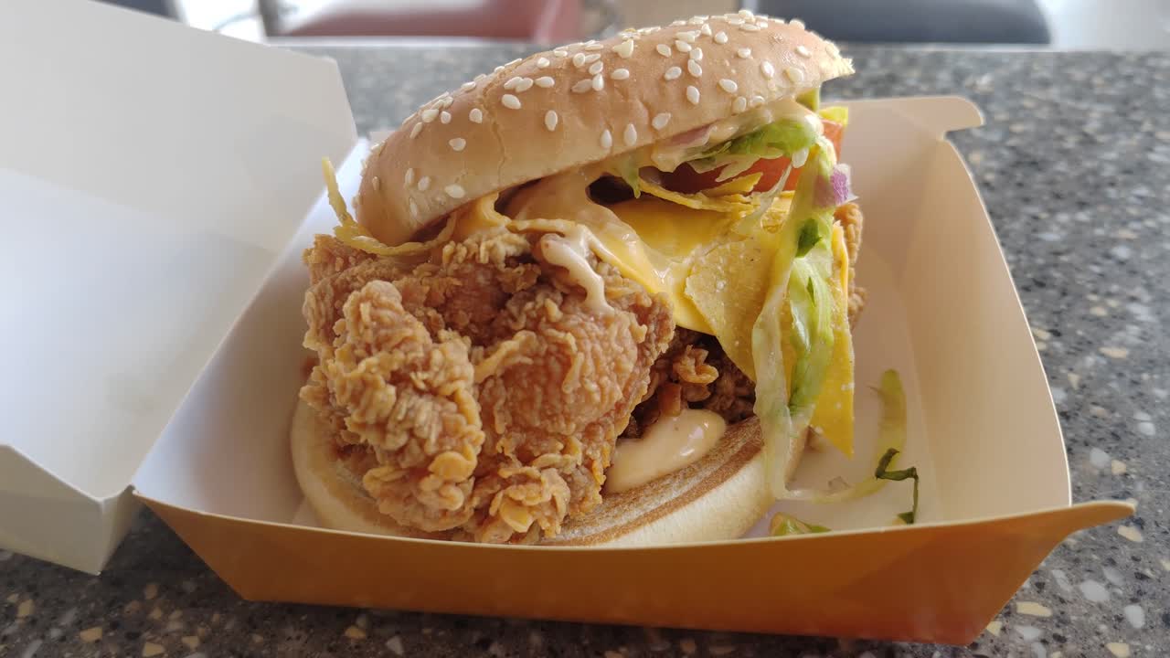 A Mexican-inspired burger with crispy chicken slice, cheese, and taco flavors served on a table inside a fast-food restaurant.
