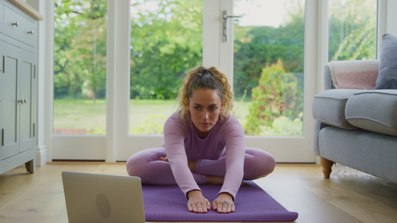 mujer joven sentada en la alfombra en casa con una computadora portátil haciendo clase de yoga en línea