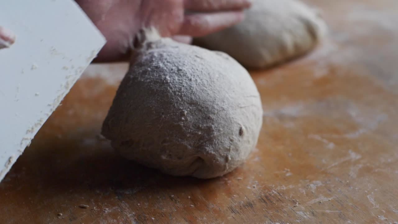 Puffy ball of dough shaped and shuffled using scraper tool on wooden kitchen tabletop, filmed as extreme closeup in slow motion shot