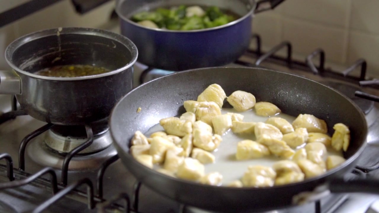Primo piano di pezzi di pollo al limone che cuociono in padella con le pinze, cibo sfrigolante sul fornello a gas, preparazione in cucina di casa con salsa e verdure sullo sfondo