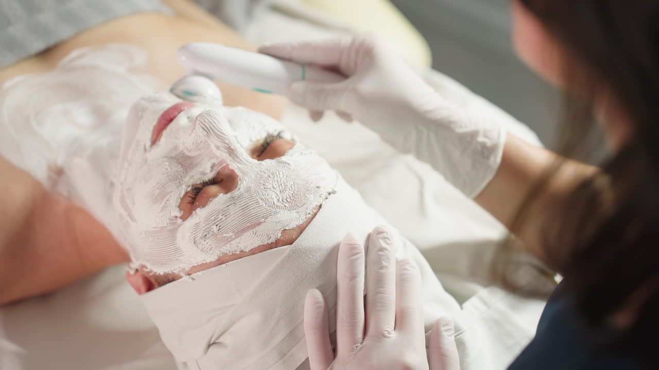 High angle view of facialist using vibrating electric facial roller with gloved hand to massage thick white cream mask on spa visitor under bright lamp for skin rejuvenation and soothing relaxation