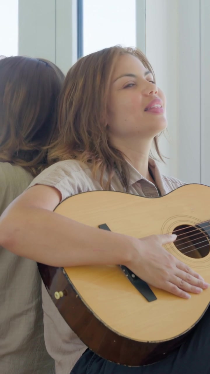 Video verticale: Donna in camicia appoggiata all'indietro che strimpella una chitarra acustica, cantando a casa vicino a una finestra con specchio.