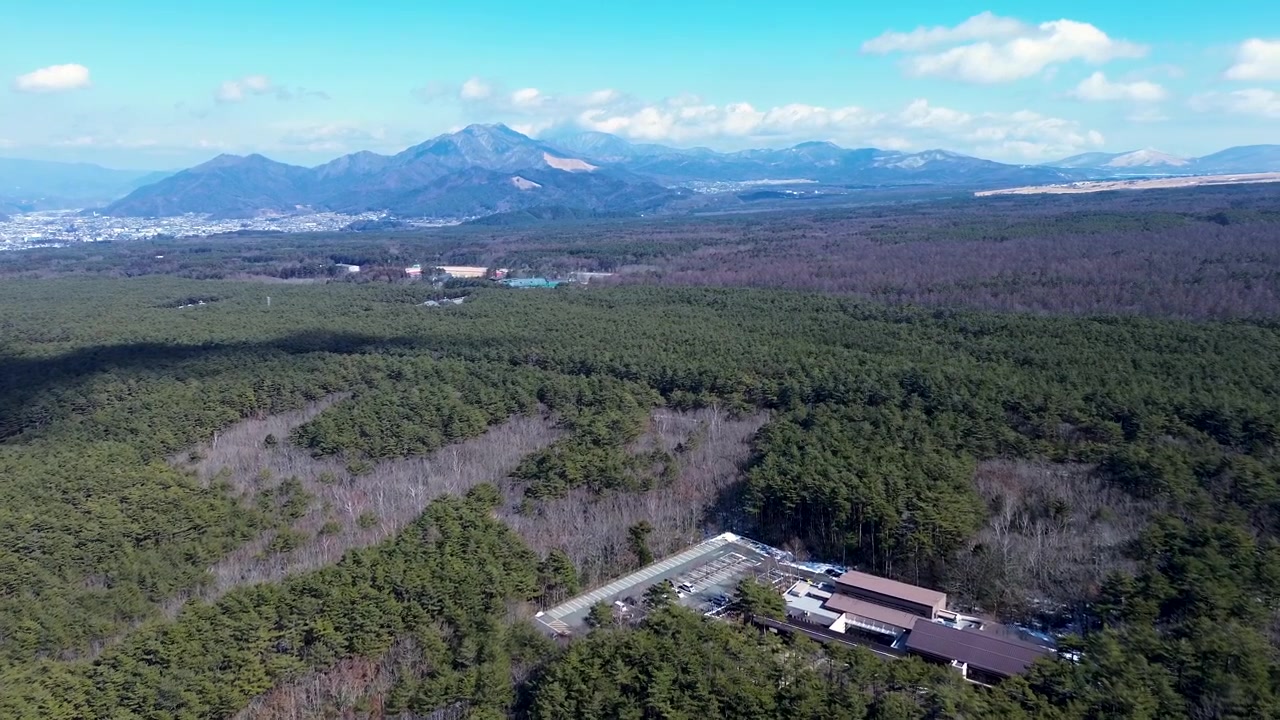 Drone-ilmakuva parkkipaikasta ja rakennuksesta, jota ympäröivät tiheät pensaikot ja metsäpuut laaksossa ja vuoristoniemekkeellä, pilvisen taivaanrannan kanssa horisontissa lähellä Kawaguchikoa, Japania, Aasiaa