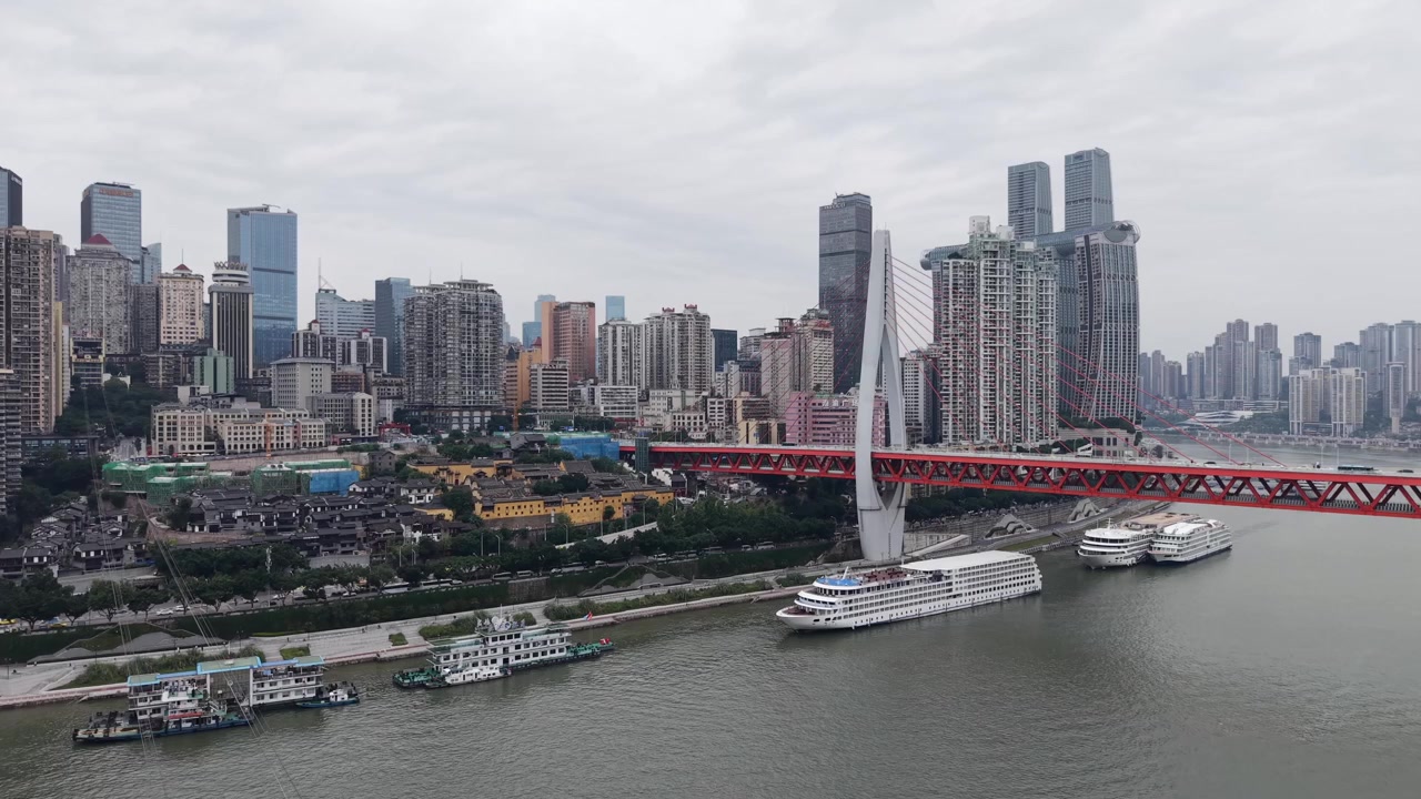 Lenta panoramica aerea sullo skyline della città di Chongqing da un ponte strallato rosso verso il denso lungofiume di grattacieli, barche sul fiume sottostante, giornata nuvolosa con foschia, Chongqing, Cina