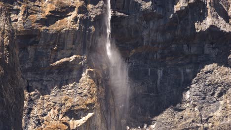 a waterfall in the wild mountains of leukerbad. the wind blows the water into the air. everything is raw and wild. a stark landscape. the rocks are illuminated by the sun