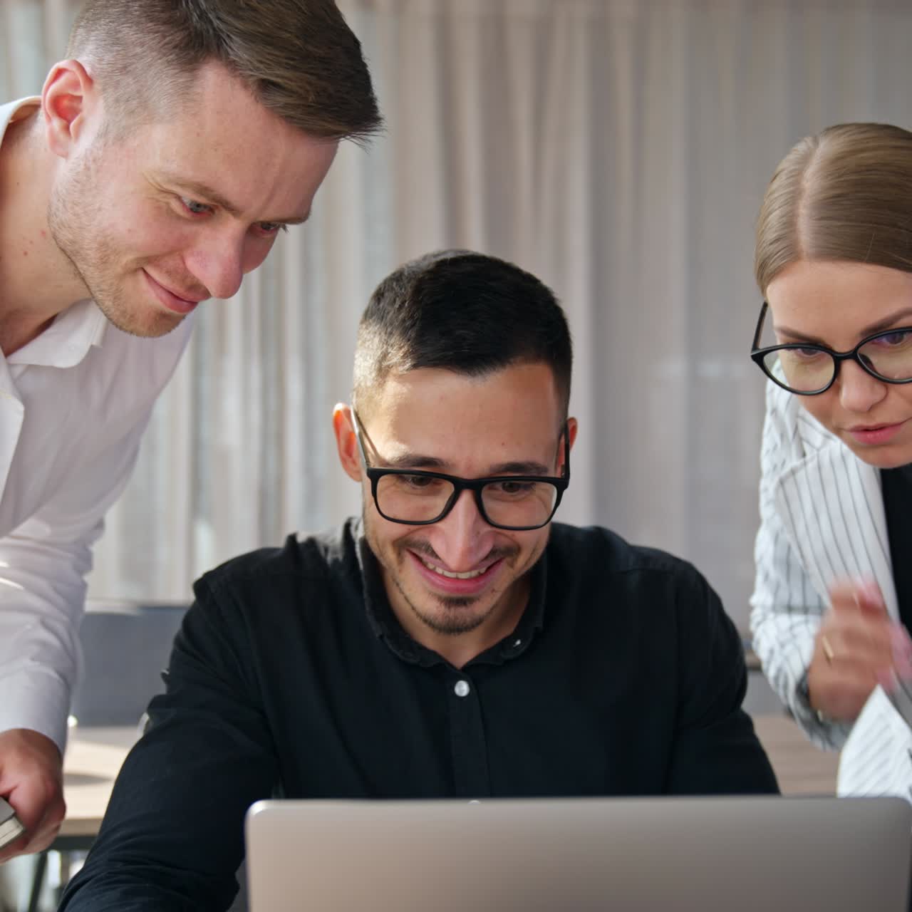 Happy smiling male wearing glasses sits in front of laptop. Two colleagues standing beside him looking intently on the screen talking