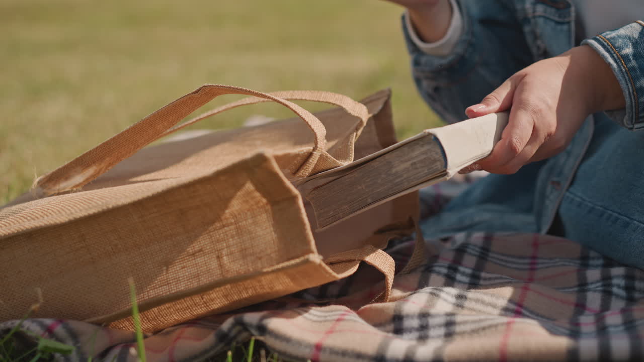 Mani che disfano una borsa di paglia su una coperta, manica di jeans visibile, erba e luce solare, texture intrecciata tattile, movimento lento e attento, atmosfera rilassata del weekend, preparazione di un momento di picnic tranquillo