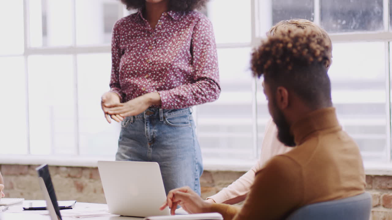 Millennial black woman stands presenting to colleagues in a meeting room, tilt shot, rack focus