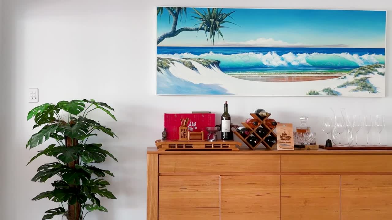 A vibrant coastal painting above a wooden sideboard featuring wine bottles and a potted plant.
