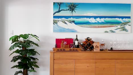 a vibrant coastal painting above a wooden sideboard featuring wine bottles and a potted plant.