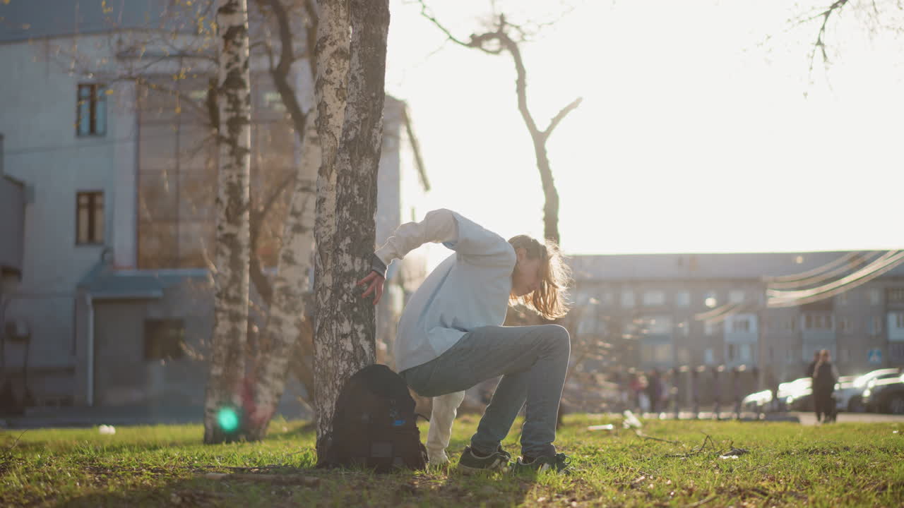 황혼녘에 공원을 걷는 젊은이, 배경에는 자작나무와 도시 건물이 보이고, 나무 줄기에 기대앉아 어깨에는 백팩을 메고 있으며, 따뜻한 렌즈 플레어와 부드러운 황금빛 조명이 평화로운 분위기를 연출해요.