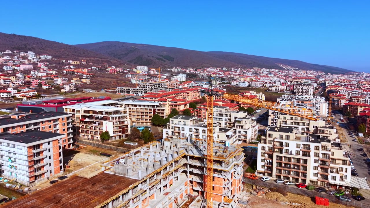 Veduta aerea di una gru da costruzione sopra una città collinare. Gru a torre arancione tra edifici con tetti rossi e un pendio montano boscoso.
