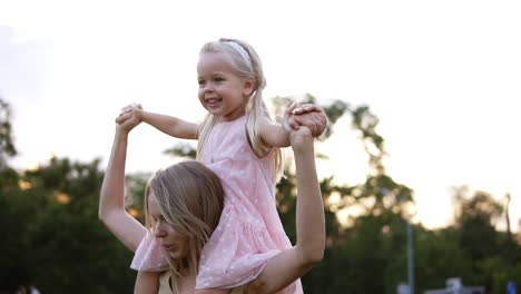 happy mother carries beautiful little daughter on her shoulders, dancing and having fun together in public, green park