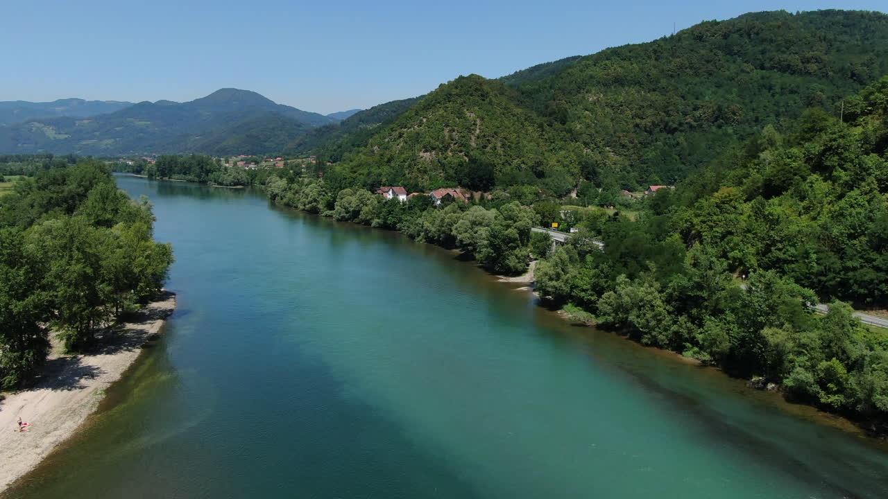 Vista de um rio largo fluindo por um vale. Há colinas verdes em cada lado. A água é clara e azul. Algumas árvores e rochas estão perto do rio