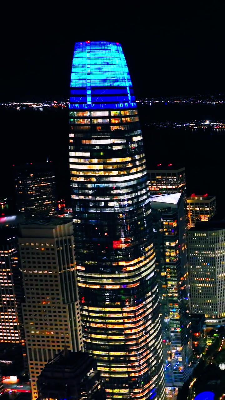 Close-up noturno da Torre Salesforce, São Francisco, Califórnia. Coroa iluminada em azul eleva-se acima de arranha-céus iluminados e da baía escura e distante. Vídeo vertical
