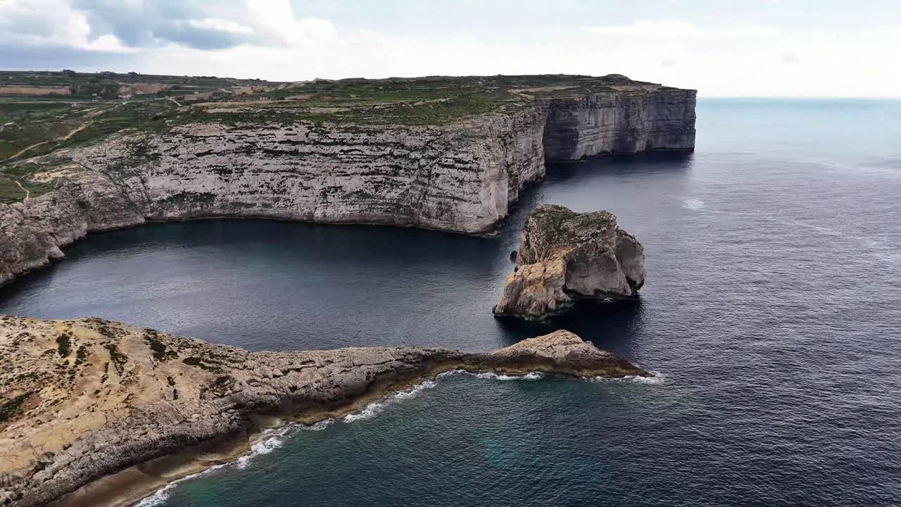 Dwejra Bay in San Lawrenz Gozo Malta shows dramatic limestone cliffs and Fungus Rock rising from Mediterranean Sea within Lower Coralline Limestone coastal system, aerial drone orbit shot