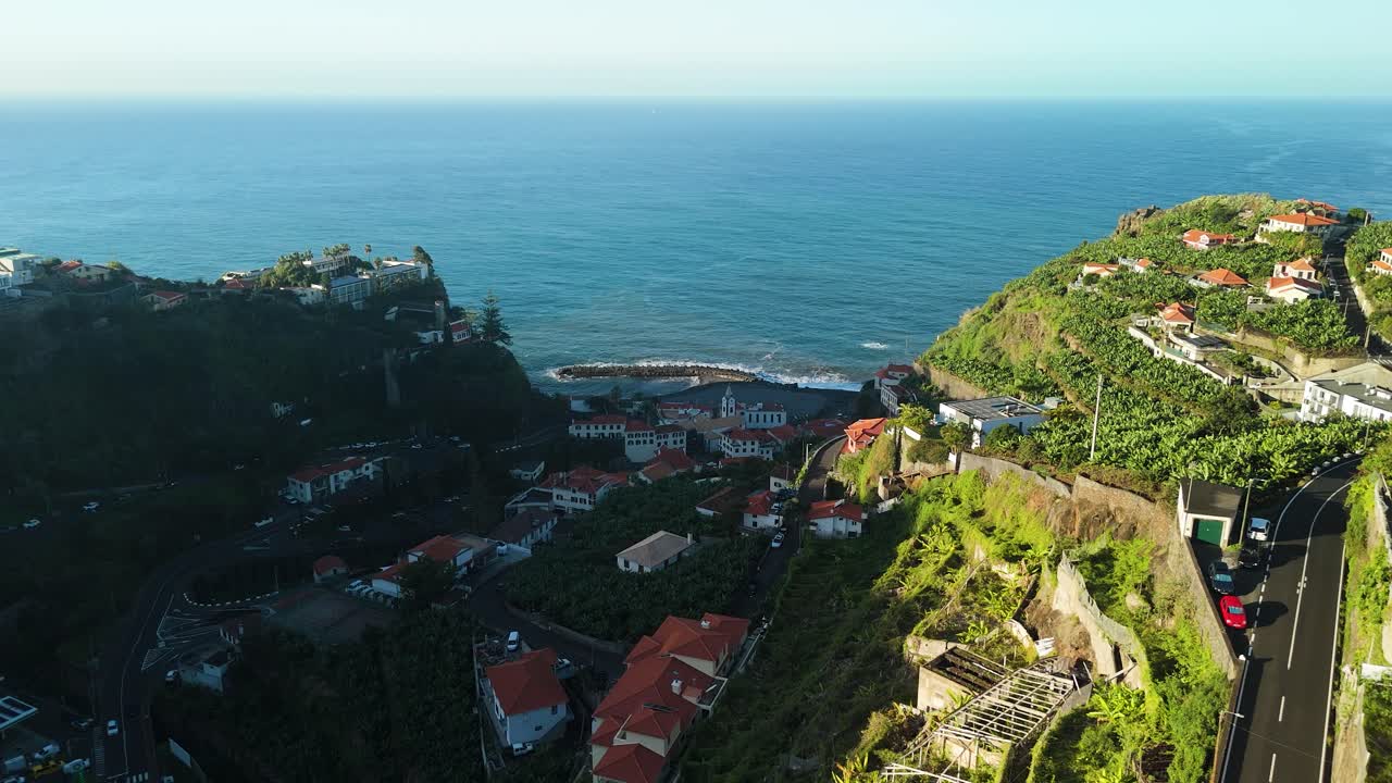 Drone zooming in on Ponta do Sol in Madeira, bathed in a sunny glow along green cliffs and a hazy blue ocean