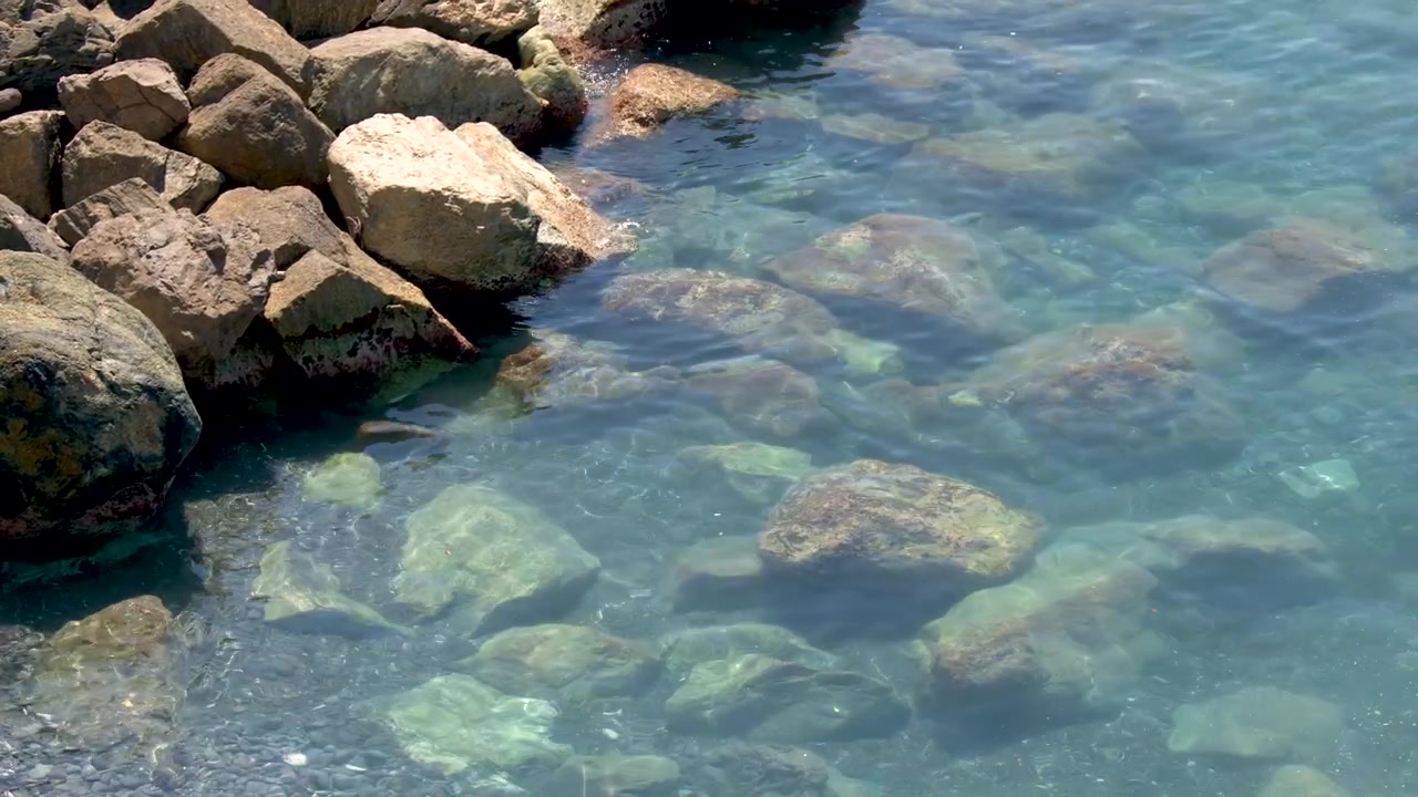Inquadratura statica dall'alto di acqua trasparente del Mar Mediterraneo che increspa dolcemente sulle rocce costiere.