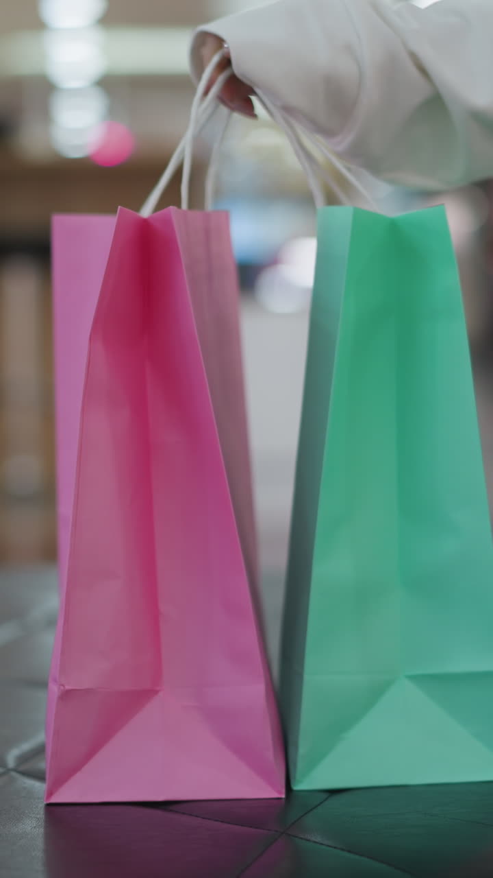 bolsas de compras rosas y menta colocadas en una superficie de baldosas, ligeramente inclinadas, con un fondo borroso del centro comercial, una mujer en vaqueros se sienta cerca en el asiento de cuero