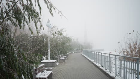 early winter cityscape with first snowfall, foggy atmosphere, snow-covered benches, light poles, and iron railings along park walkway, framed by frosted trees and muted, serene surroundings