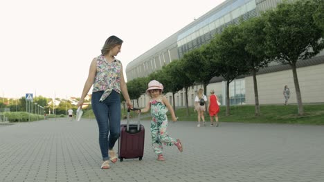 mother and daughter walking outdoors to airport. woman carrying suitcase bag. child and mom vacation