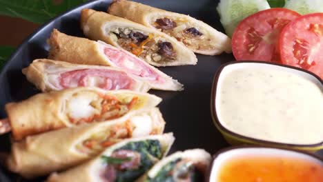 delicious spring rolls with dipping sauce and sliced tomatoes and cucumber on a black spinning plate close up