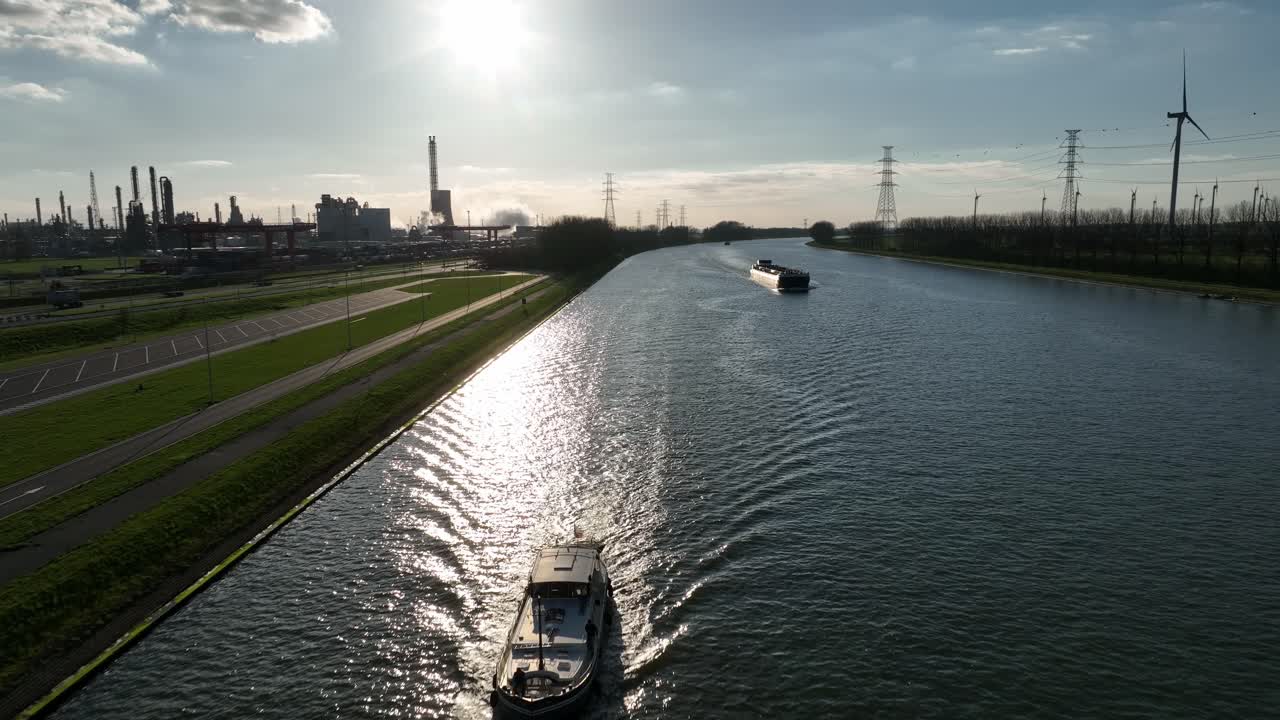 Zoom out aerial shot of Antwerp canal with boats sailing under afternoon sun, surrounded by factories, pipelines, and industrial facilities