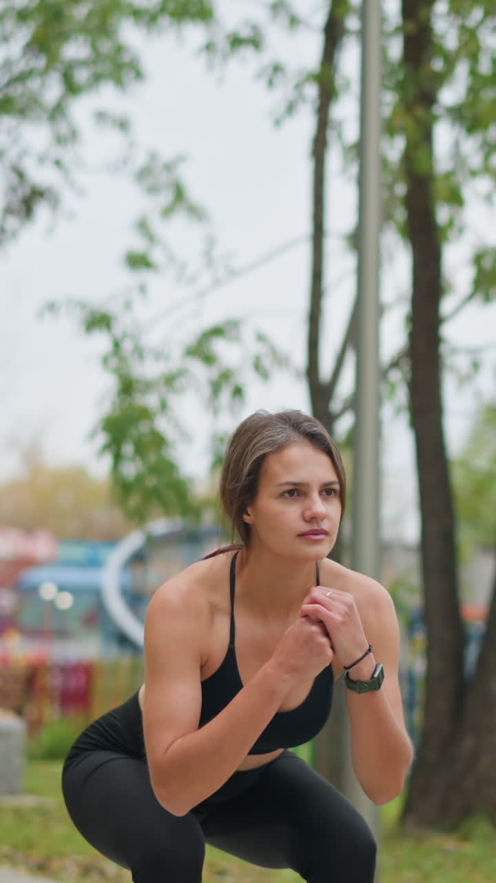 Young lady squatting down in game center outdoors with a blurred background featuring trees and colorful playground equipment, emphasizing fitness, strength, and outdoor exercise