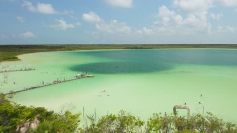 Backwards-fly-above-amazing-colourful-lake-in-nature.-People-swimming-and-taking-rest-in-sunny-day.-Kaan-Luum-lagoon,-Tulum,-Yucatan,-Mexico