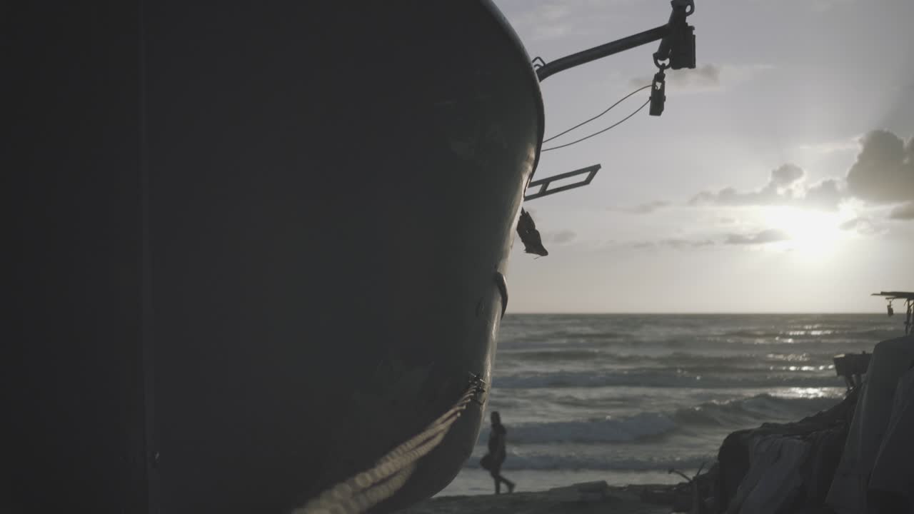 Uomo che cammina lungo la riva da dietro una barca mentre il sole tramonta nel mare - Termina con un flare e un movimento di panoramica da sinistra a destra