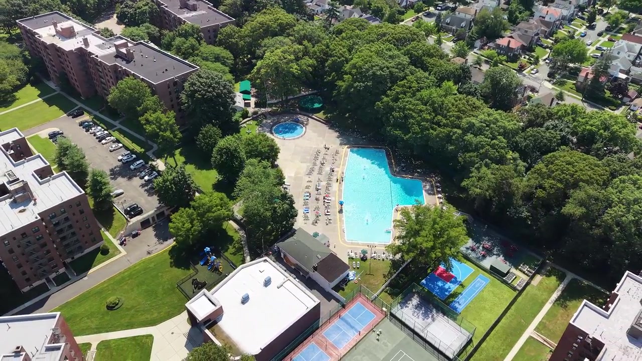 Flushing queens ny drone's perspective flying over a large swimming pool in a residential complex with people relaxing and sunbathing on lounge chairs during a sunny summer day in a suburban neighborh
