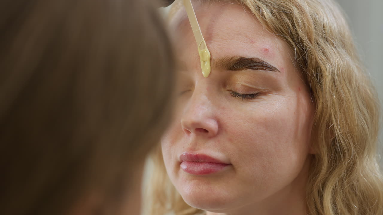 Close up of beautician applying warm wax near client eyebrow using wooden spatula during grooming procedure, showcasing precision and care in personal beauty service inside professional salon