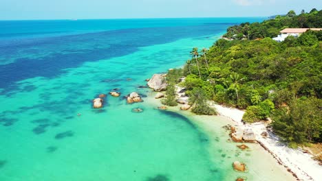 Tropical-island-with-palm-trees-hanging-over-white-sandy-beach-and-limestone-cliffs-washed-by-turquoise-lagoon,-Thailand