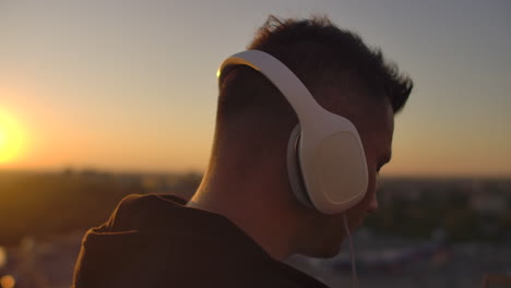 Rear-view-of-a-man-in-headphones-listening-to-music-and-working-on-the-roof-of-a-building-at-sunset-with-a-view-of-the-city-from-a-height.-Roof-of-a-skyscraper-at-sunset.