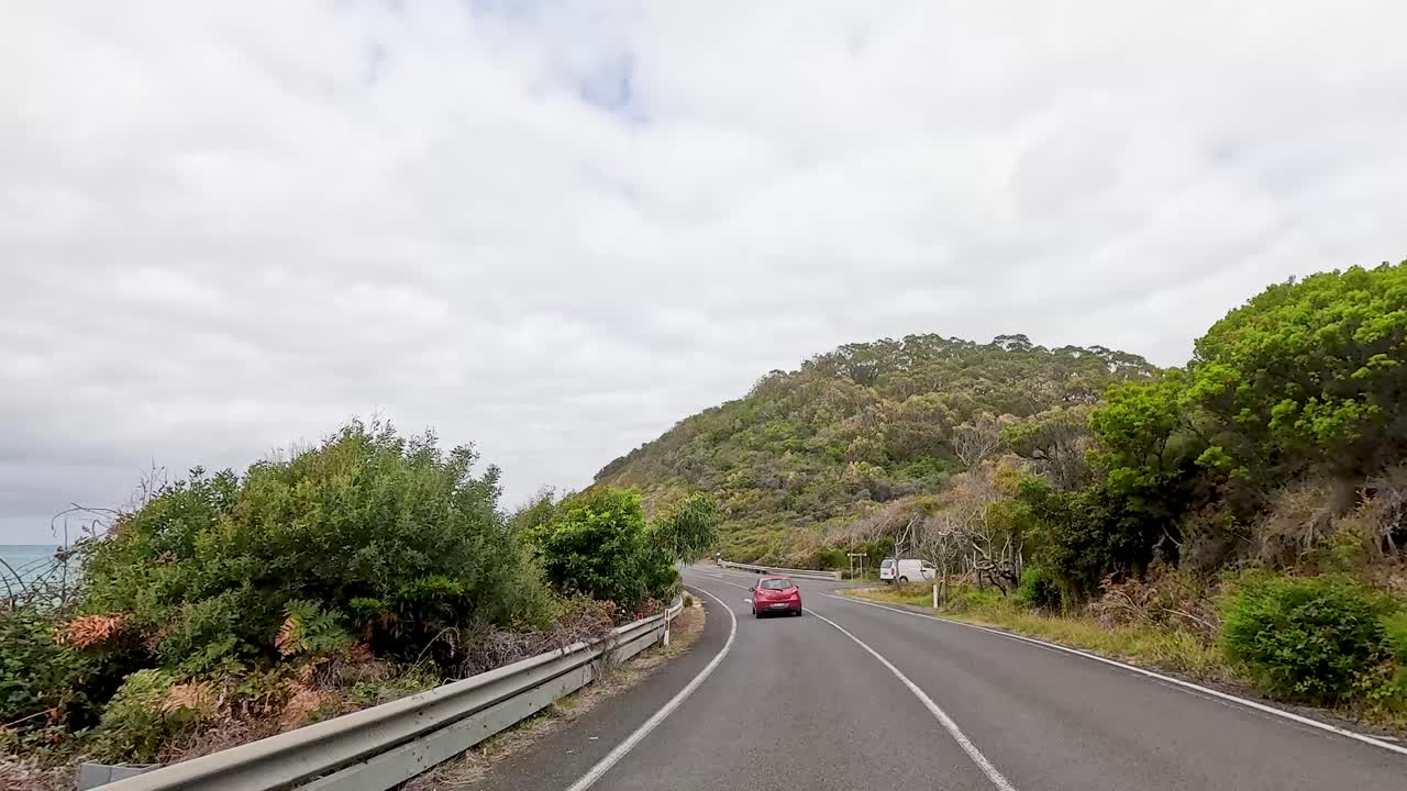 A car travels along the picturesque Great Ocean Road, surrounded by lush greenery and coastal views under a cloudy sky