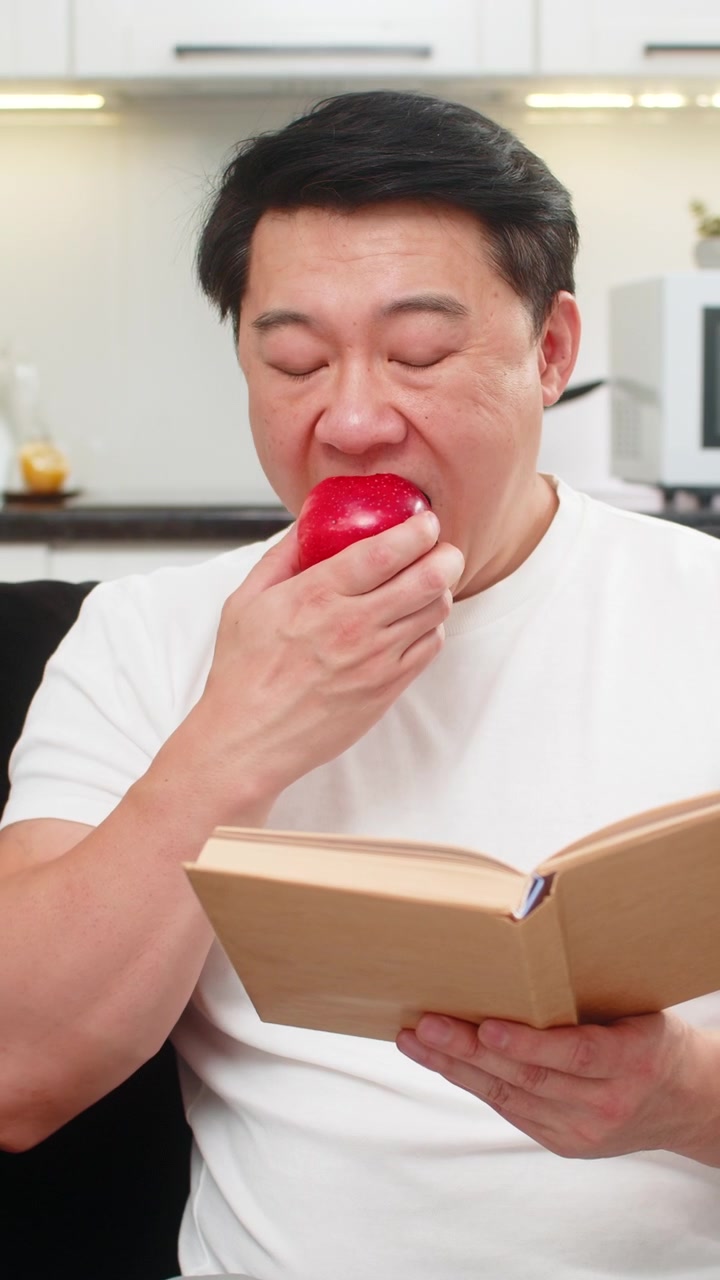 赤いリンゴをかじり、ジューシーな味を楽しんでいるアジア人男性が、本で短い健康のヒントを穏やかな笑顔で読んでいる。