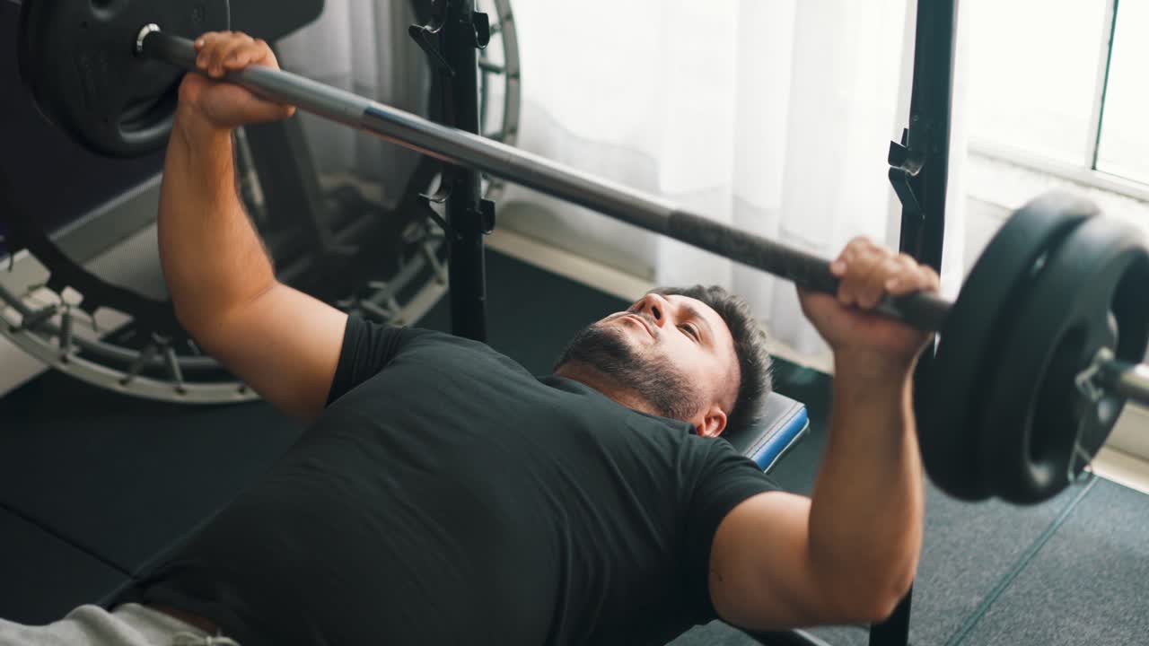 Bench press'te ağırlık kaldıran genç adam, arkasında bir trambolinle.