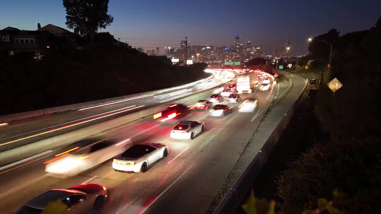 Una vista de larga exposición del tráfico vespertino creando hermosas estelas de luz en una autopista concurrida, con el icónico horizonte de San Francisco brillando al fondo mientras el día transita a la noche