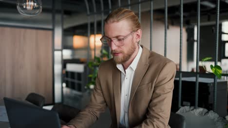 A-blond-guy-in-glasses-with-a-beard-in-a-light-brown-suit-works-at-a-laptop-and-then-looks-at-the-camera-and-smiles.-Portrait-of-a-modern-office-worker