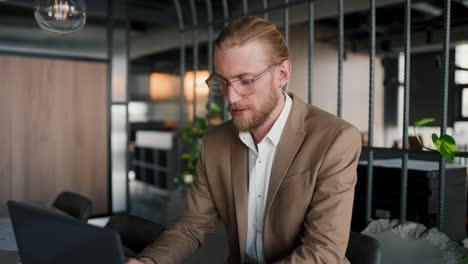 Un-Chico-Rubio-Con-Gafas-Y-Barba-Con-Un-Traje-Marrón-Claro-Trabaja-En-Una-Computadora-Portátil-Y-Luego-Mira-La-Cámara-Y-Sonríe.-Retrato-De-Un-Oficinista-Moderno