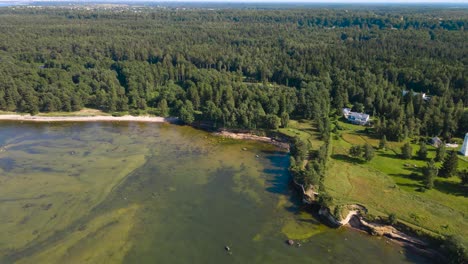 aerial view panning slowly over a sunny summer rocky beach seaside shoreline with a sandstone cliff bank where a wooden white lighthouse is placed in the middle of a large forest in the horizon