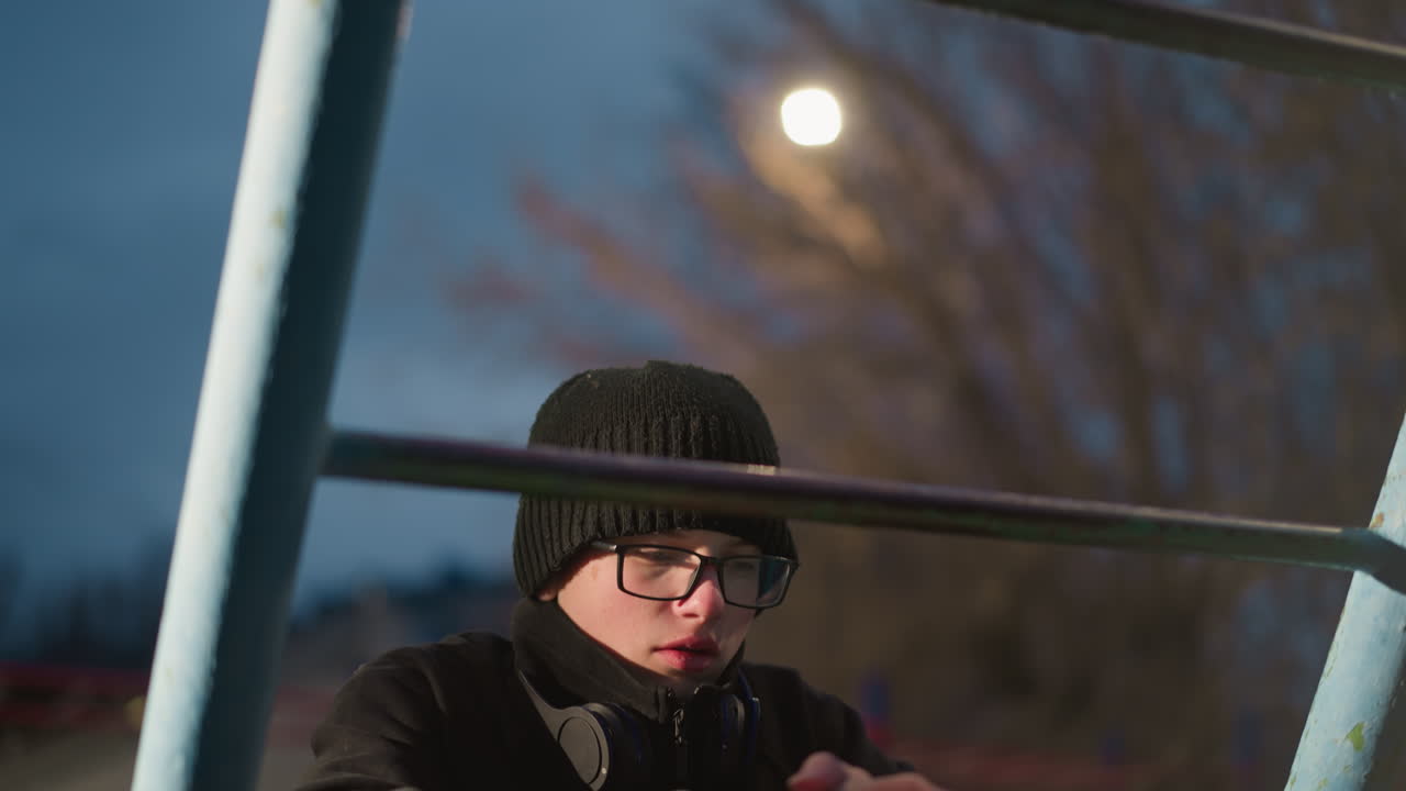 крупный план молодого мальчика в очках и черной куртке, опирающегося на железный стержень с руками, с размытыми огнями и голыми деревьями на заднем плане