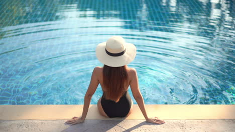 a healthy, fit sexy woman sits on the edge of a swimming pool back to the camera as she admires the view