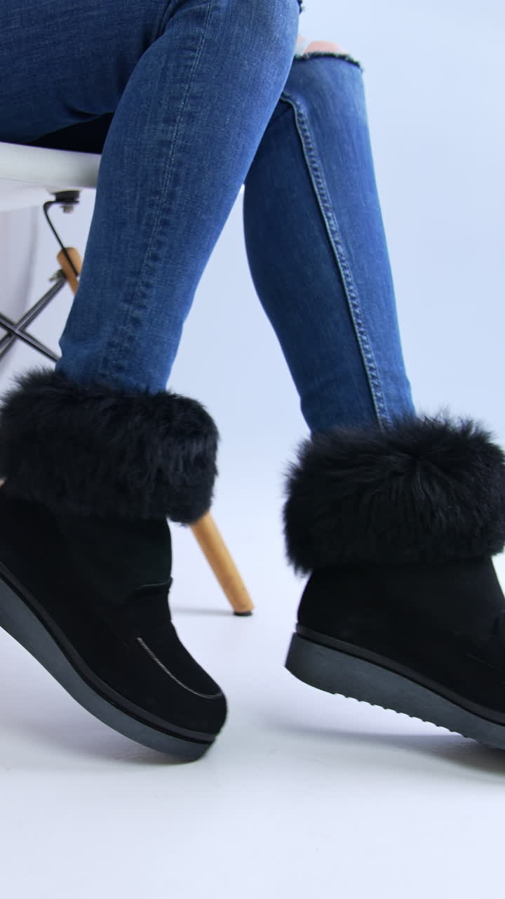 Warm comfortable low suede black boots with fur on top. Model in jeans sits on the chair showing her footwear. Close up. Vertical video