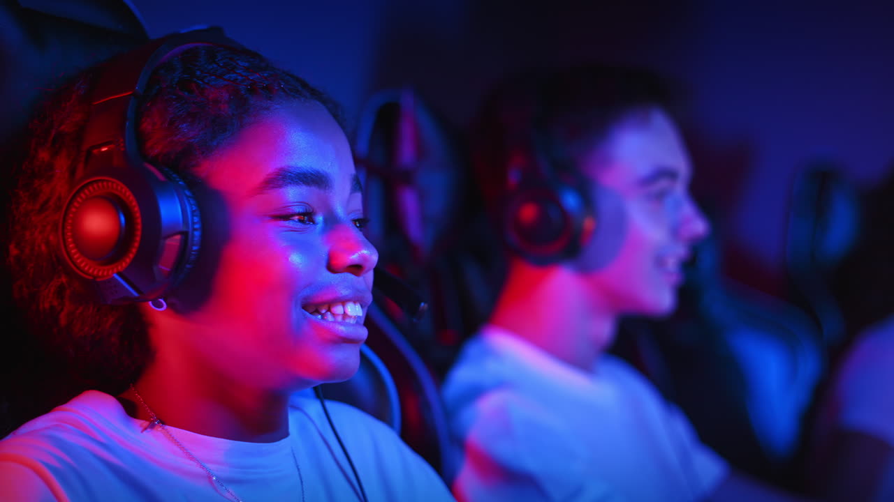 White boy and black girl teens in headsets playing video games in video game club with blue and red illumination, talking in voice chat. Slow motion