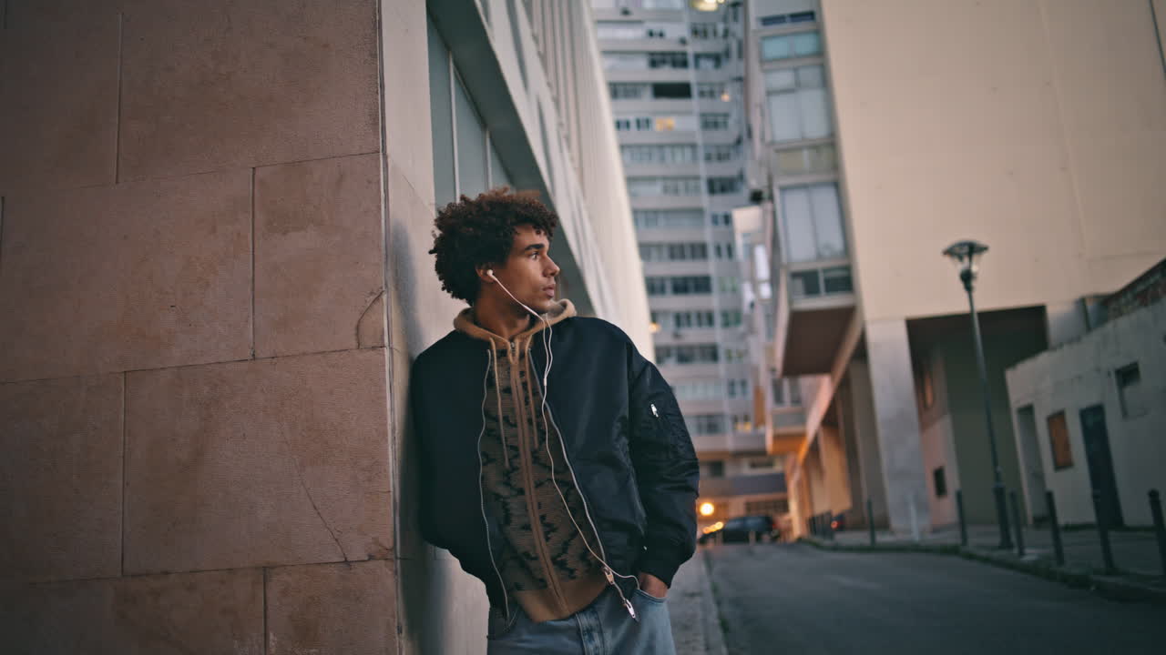 곱슬한 십대가 머리 위에 서서 헤드폰을 착용한 도시 황혼. 음악을 듣는 남자