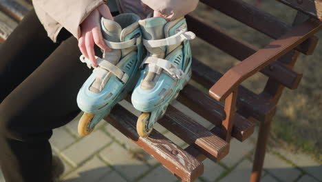 close-up of a person holding rollerblades with both hands while sitting down on a park bench. the wooden bench is visible with signs of wear, and the person's rollerblades are placed beside them