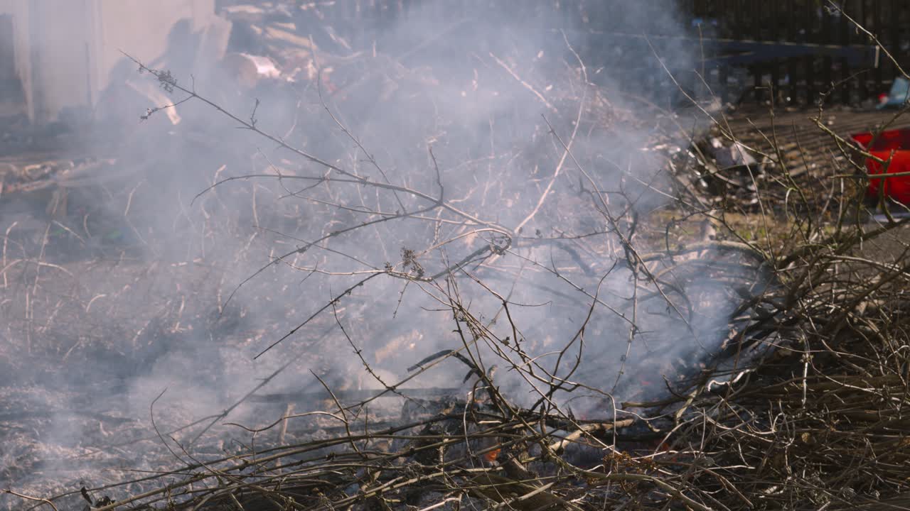 Smoldering Bonfire of Piles of Wooden Sticks and Twigs with Garbage Pile in Background